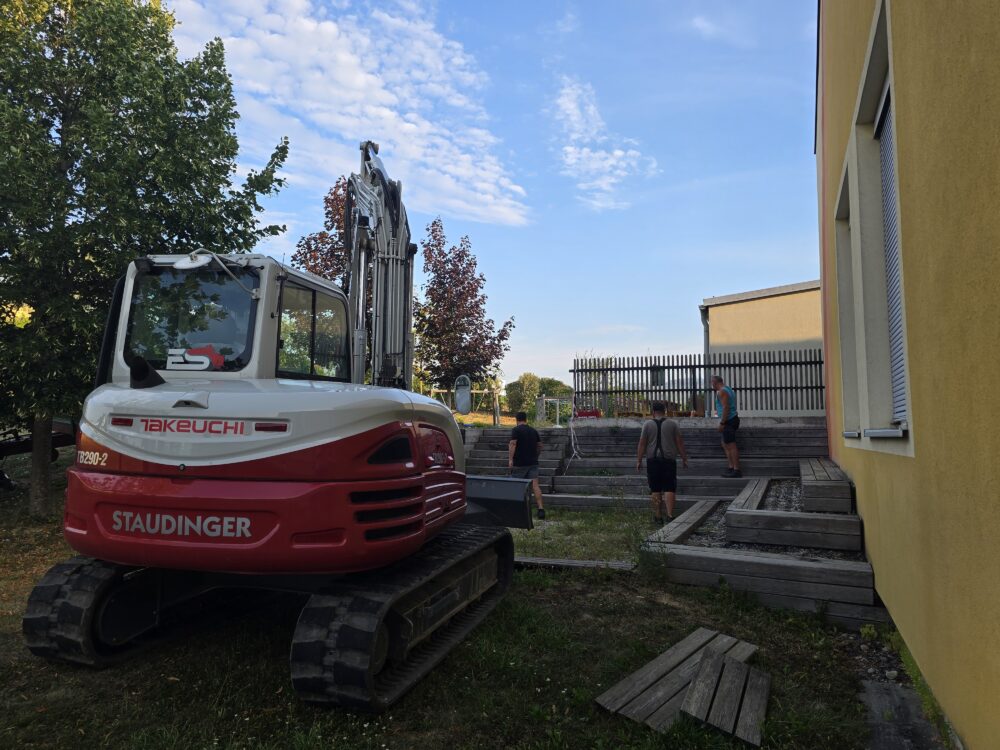 Bagger der Marke Takeuchi TB290-2 der Firma Staudinger steht neben einem Gebäude mit Holzstufen